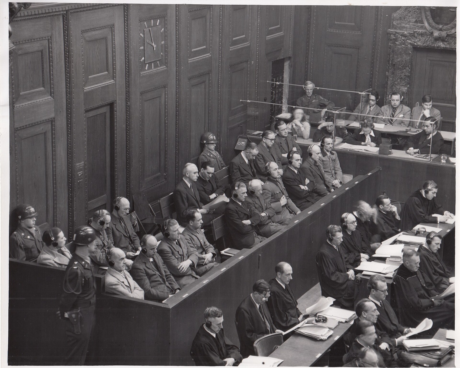 Two rows of men in suits sit in a wooden dock wearing translation headphones, with helmeted guards standing behind them.