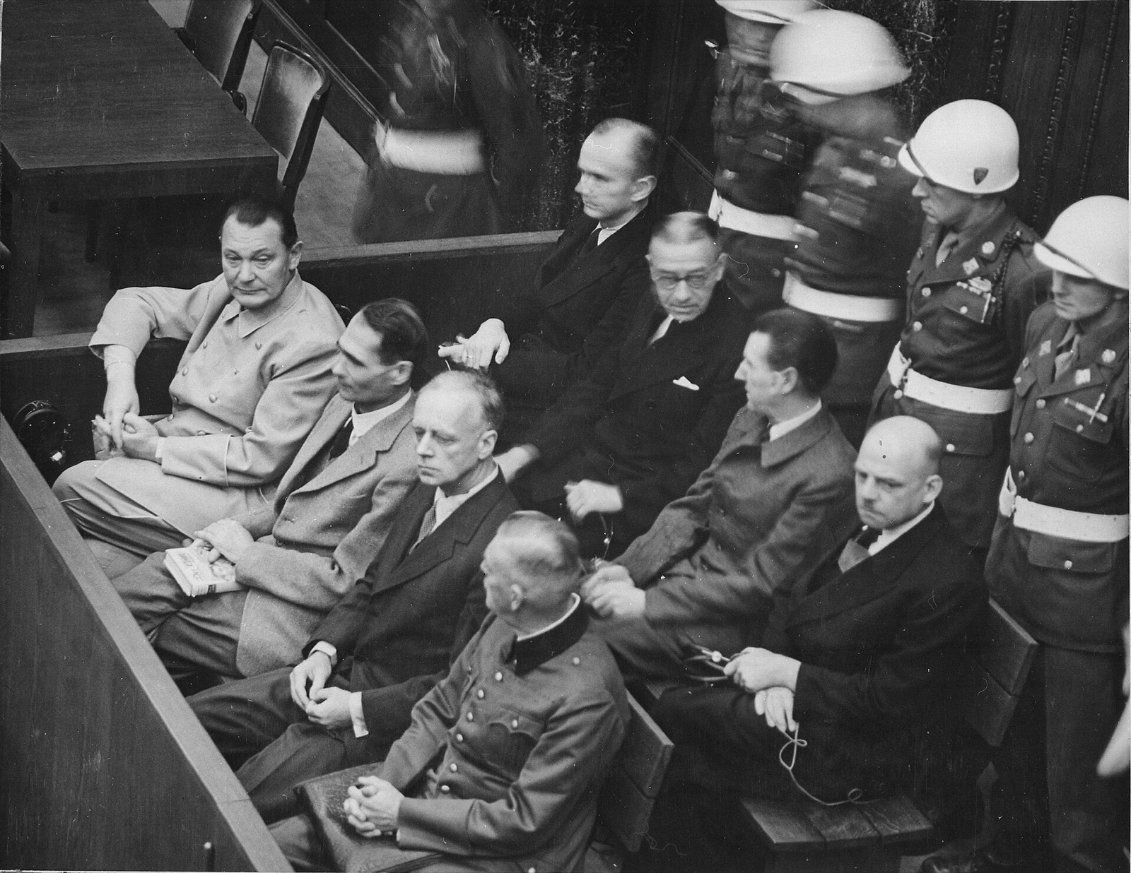 Two rows of men seated in a wooden dock, with military police standing behind them along the wall.