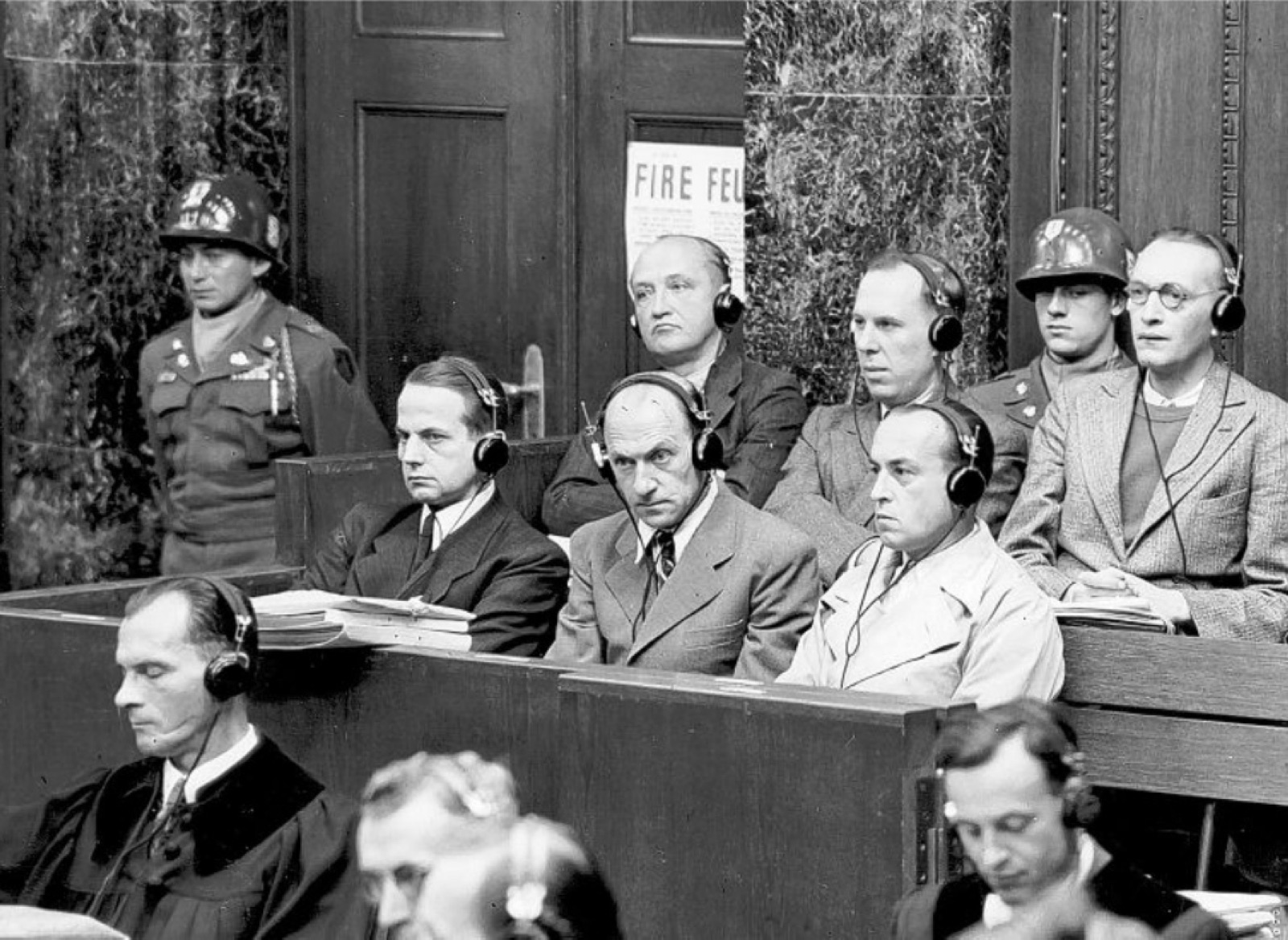 Six men in suits sit in two rows in a courtroom dock, several wearing translation headphones.