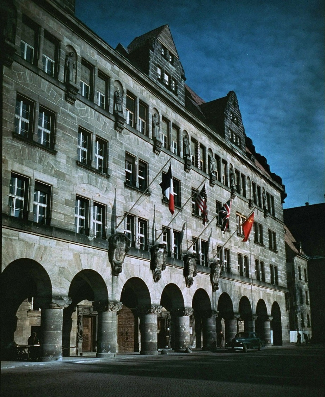 Four national flags hanging vertically side by side from the stone facade of a large public building.