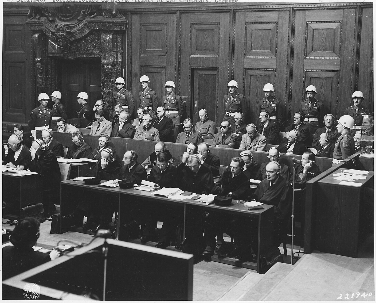 Two rows of men in suits and uniforms seated in a wooden dock, helmeted American military police standing shoulder to shoulder behind and beside them, and the defense counsel benches in the foreground.