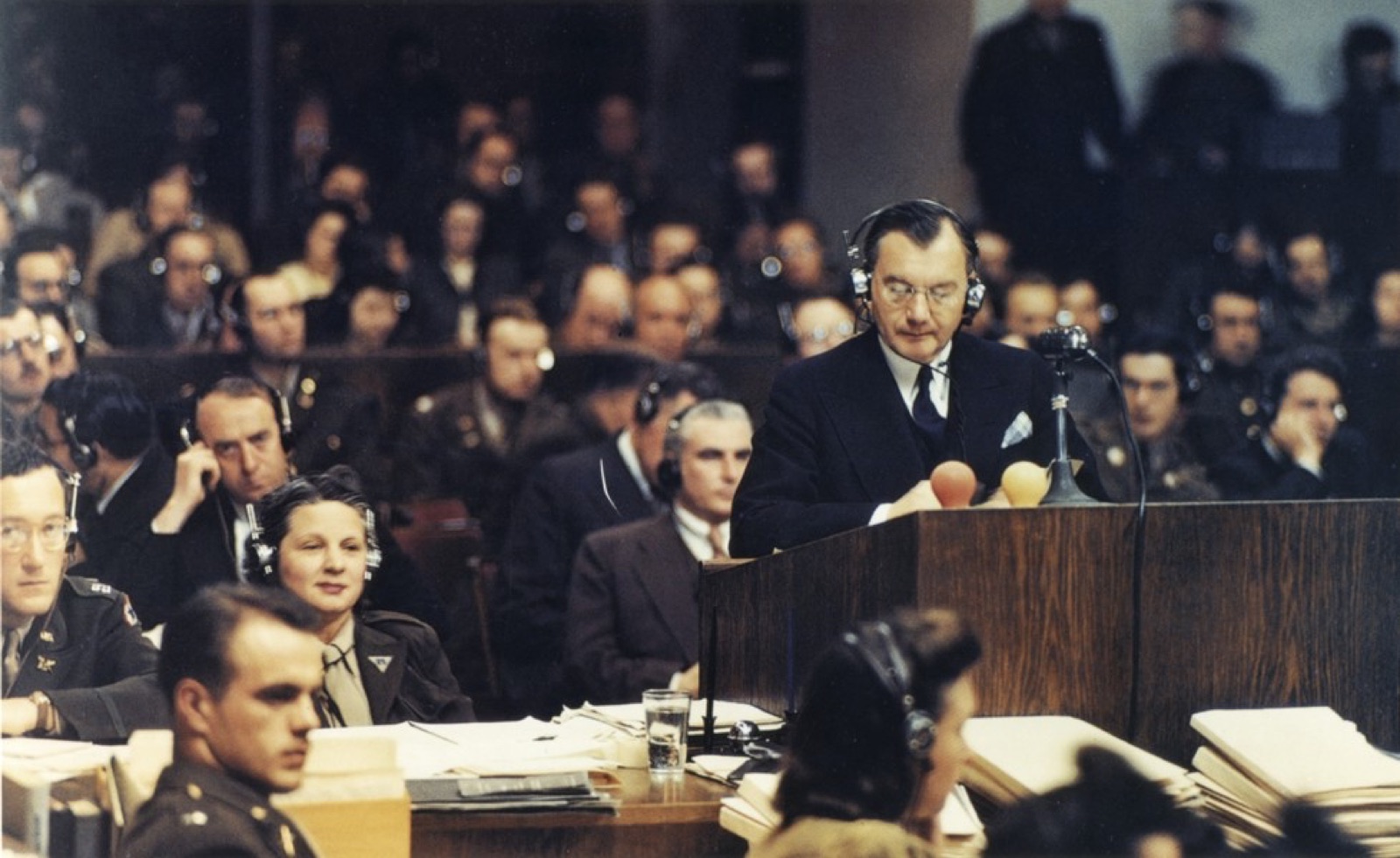A man in a dark suit standing at a lectern addressing a courtroom, photographed in color from the side.