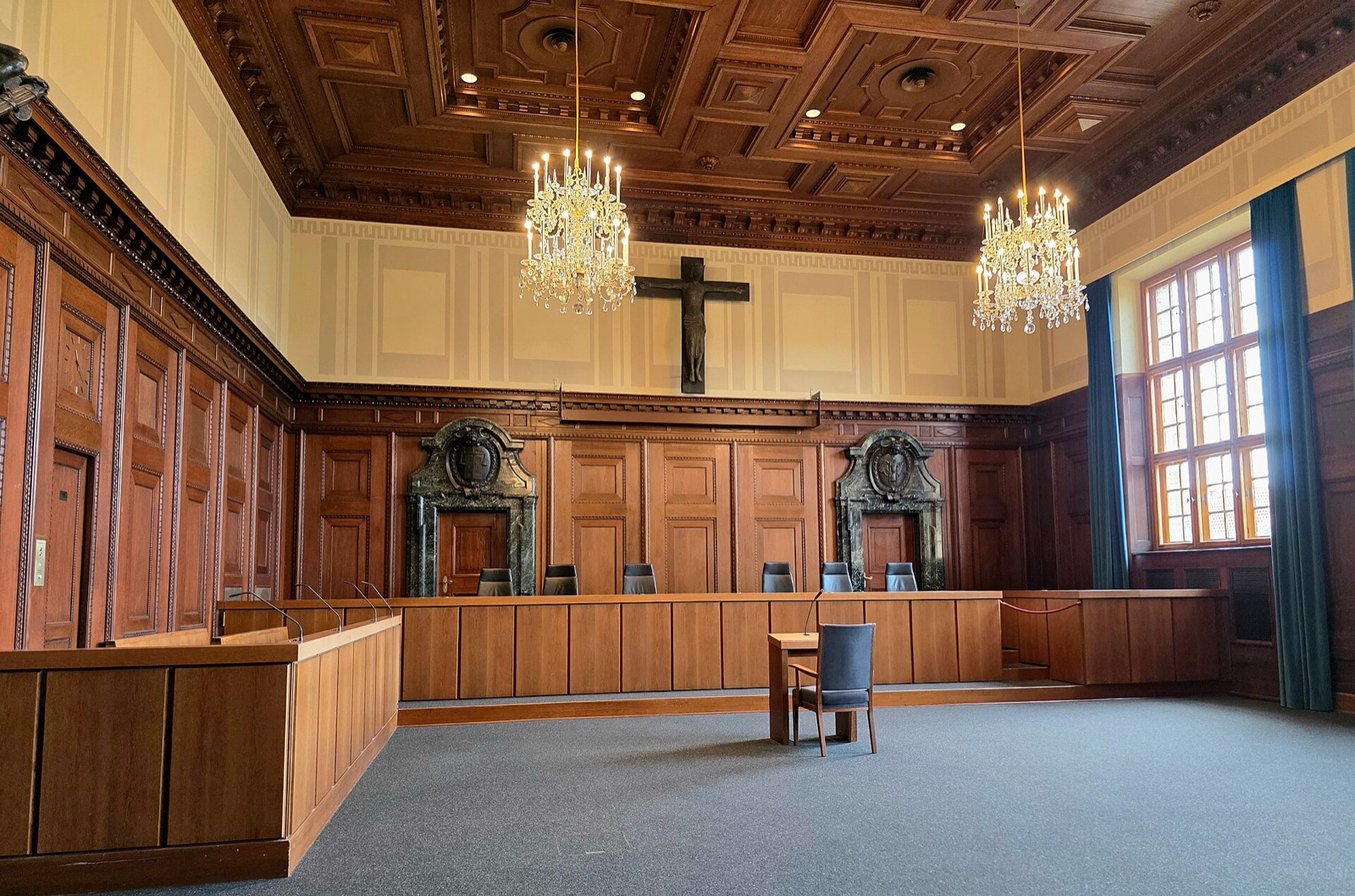A wood-paneled courtroom photographed empty from the rear, with rows of benches, tall windows along one wall and a raised judges' bench at the far end.