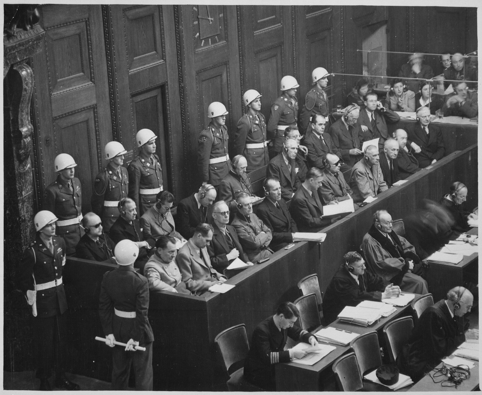 Overhead view looking down into a wooden dock holding two rows of men in suits and uniforms, with helmeted military police standing along the wall behind them.