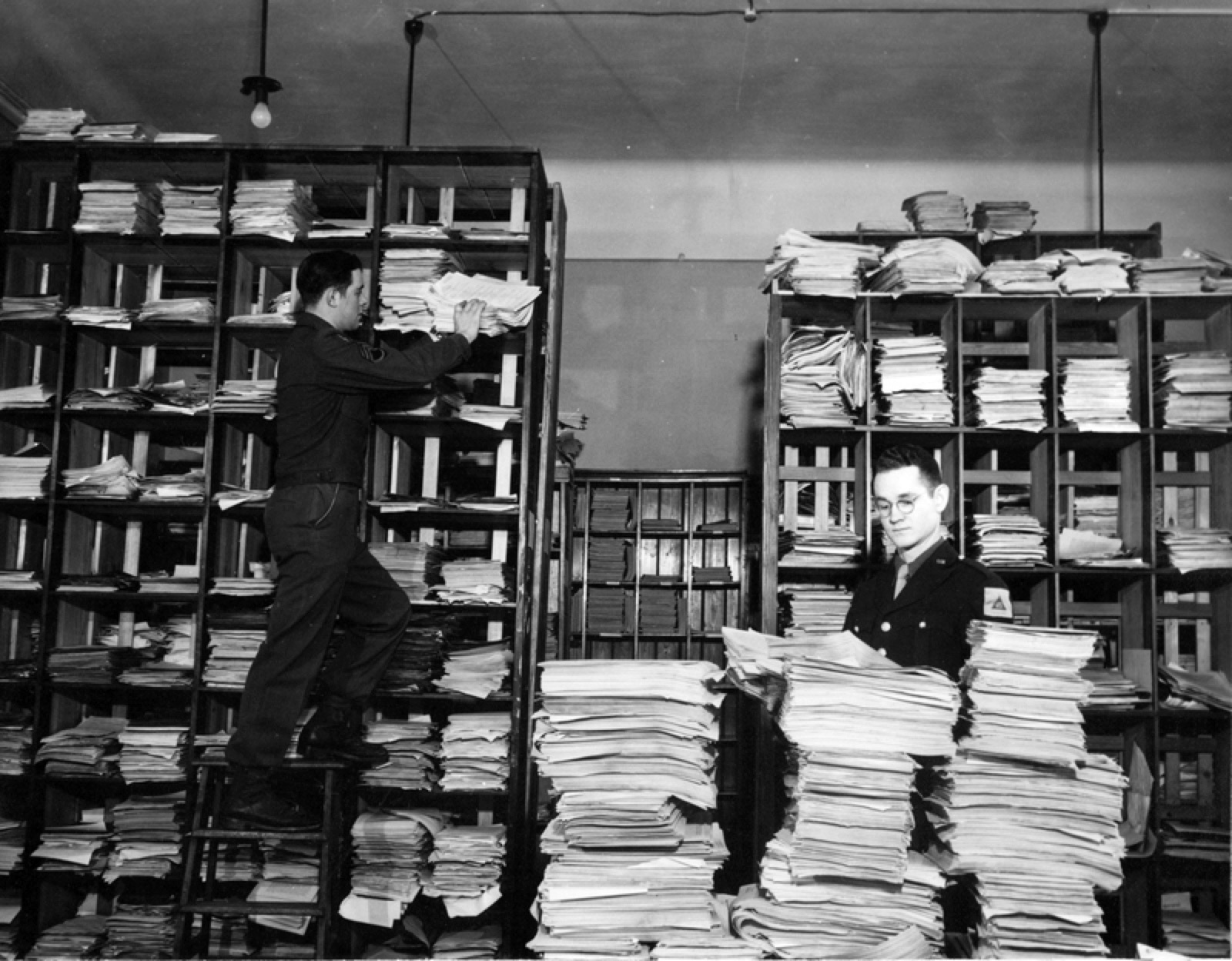 A large office room filled with rows of desks, filing cabinets and stacked document boxes, with staff working at tables.