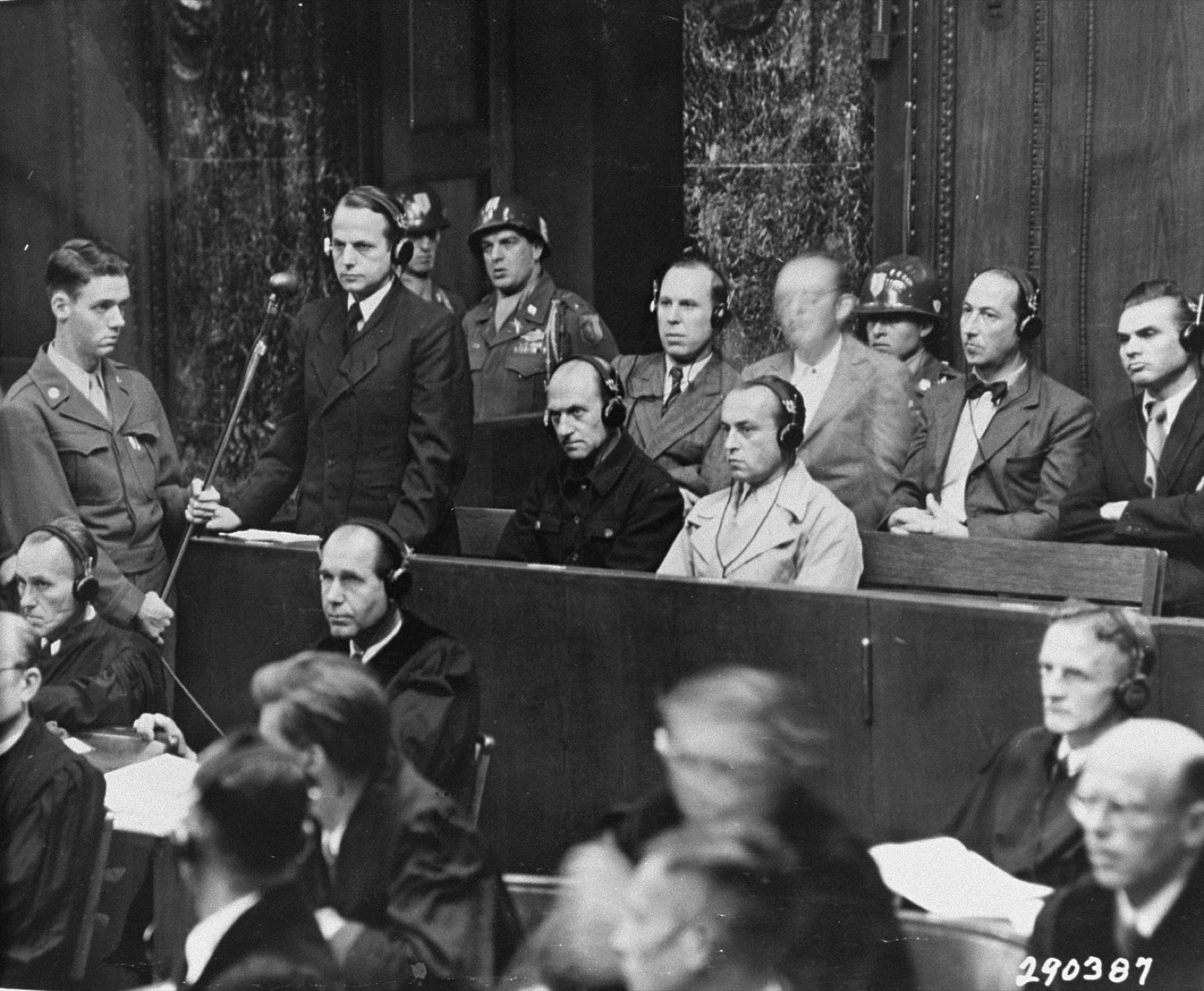 A man in a dark suit stands at a microphone in a courtroom dock, a helmeted guard immediately behind him.