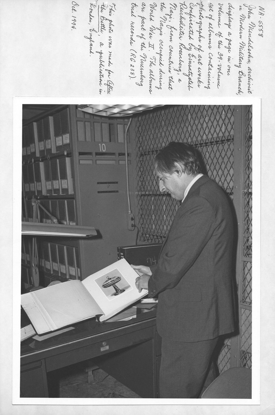 A man in a suit stands at a desk inside a wire-mesh records cage, turning an oversized album that lies open to a mounted photograph of a single object. Shelves of binders fill the wall behind him.