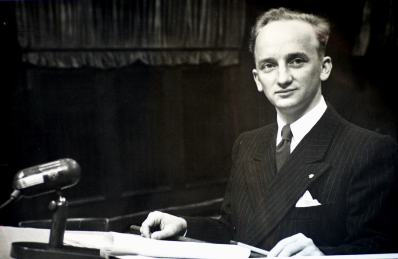 A young man in a civilian suit stands at a courtroom table, documents spread in front of him.