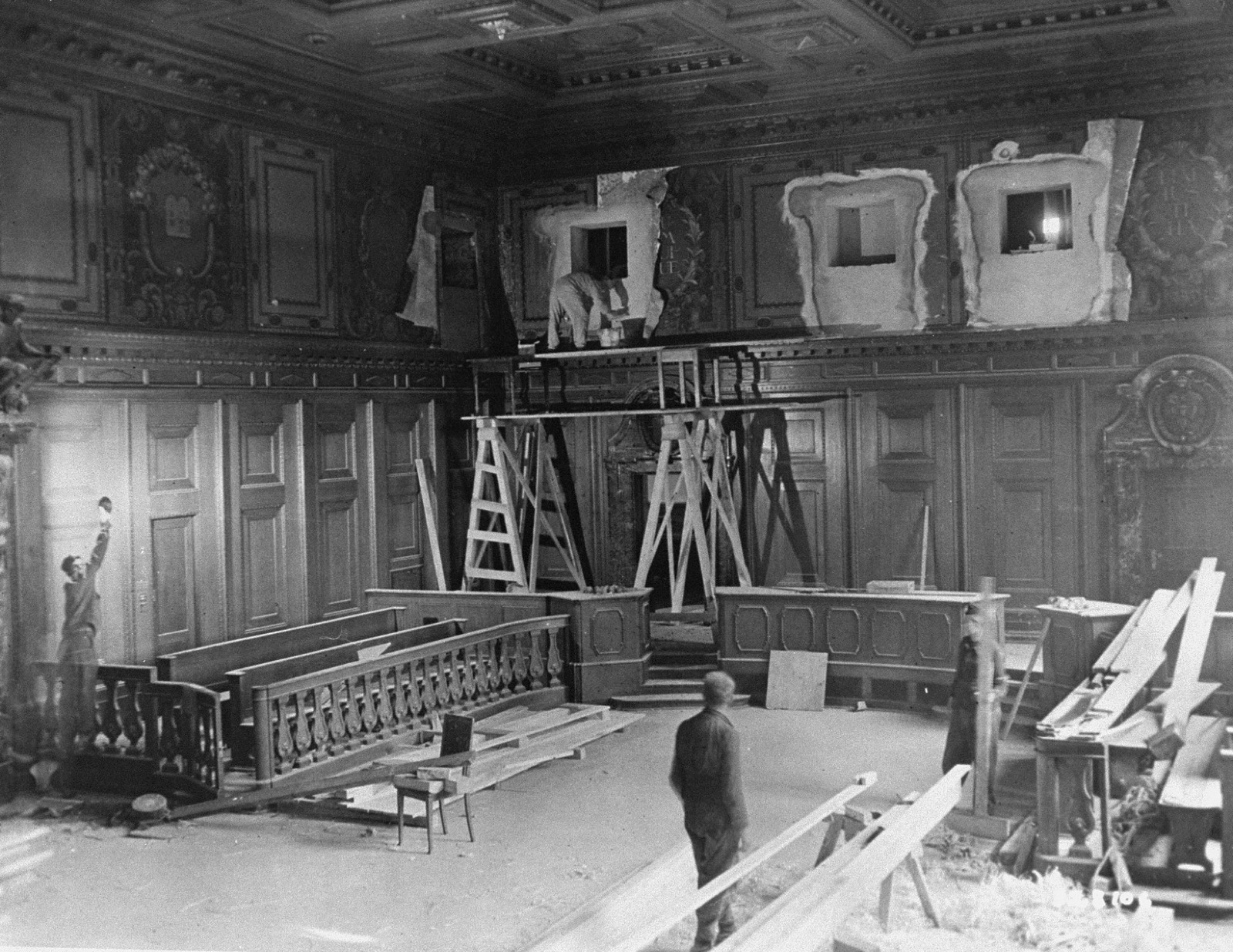 Workmen on scaffolding and ladders inside a large courtroom under renovation, with bare walls and construction materials on the floor.