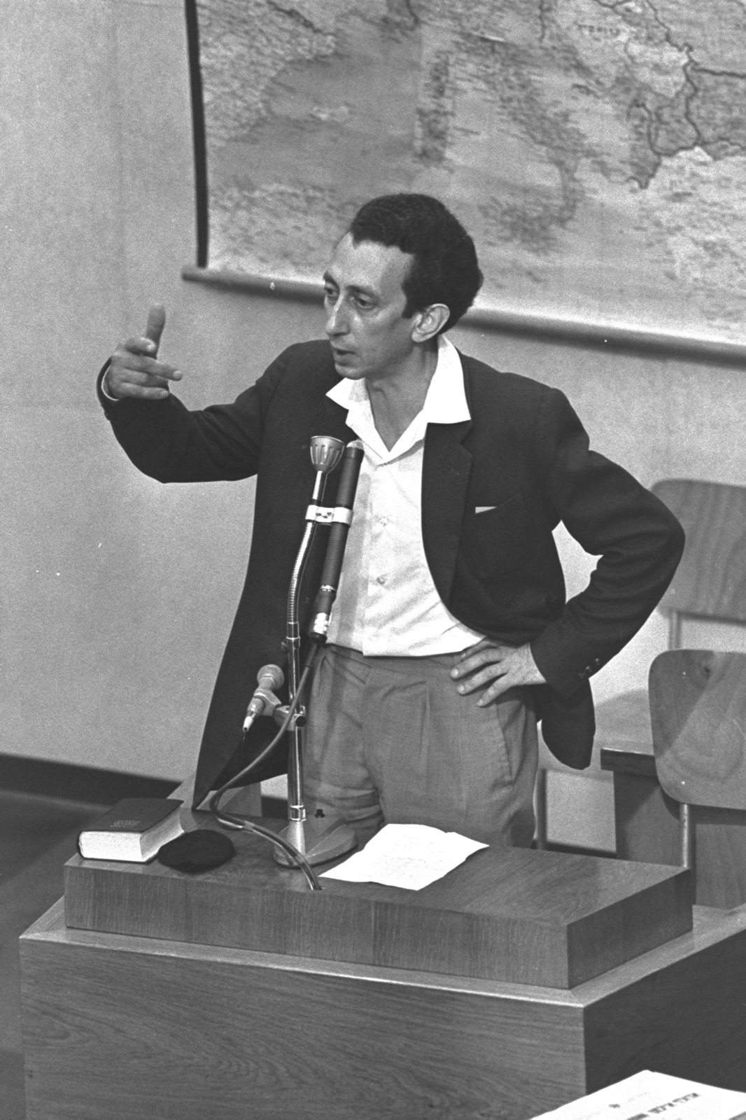 A dark-haired man in a jacket and open-necked shirt speaking at a witness stand, leaning slightly forward toward a microphone.