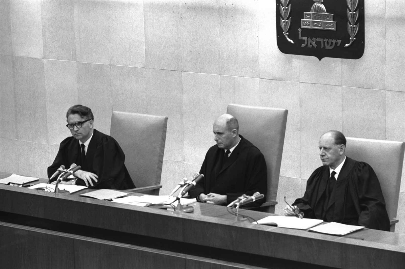 Three men in dark judicial robes seated side by side at a raised bench, papers in front of them, a plain paneled wall behind.