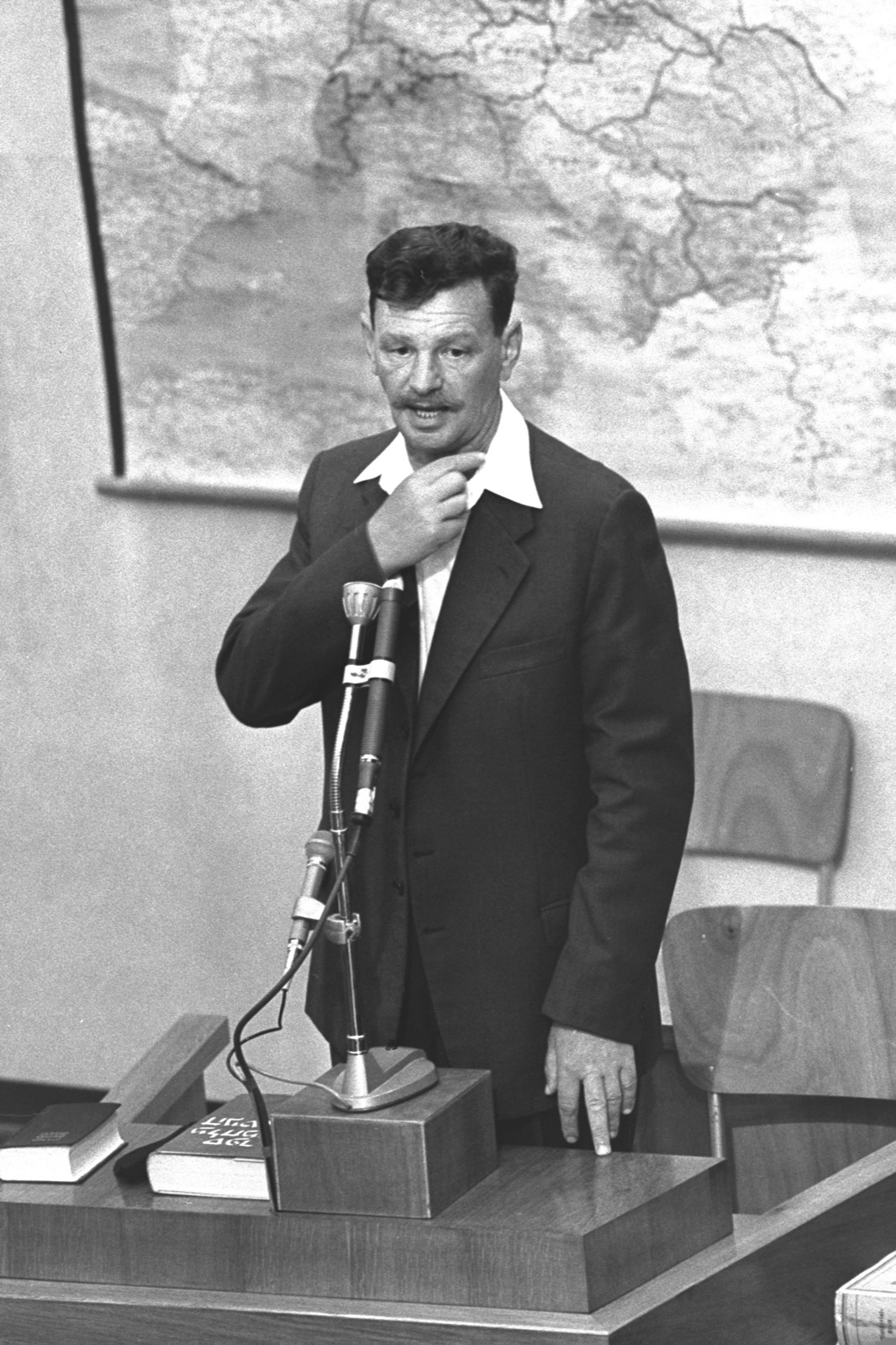 A man in a dark suit stands at a witness stand facing the court, papers on the ledge in front of him.